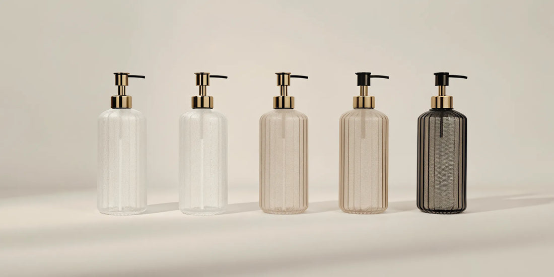 Five stylish soap pump bottles in various materials arranged by a modern sink.