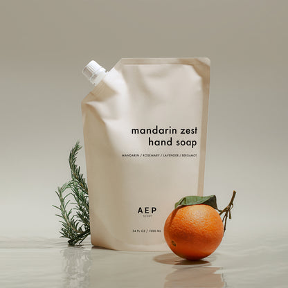 AEP Scent Mandarin Zest hand soap refill pouch made in California, featuring notes of mandarin, rosemary, lavender, and bergamot, with a fresh mandarin and rosemary sprig beside it on a beige background.