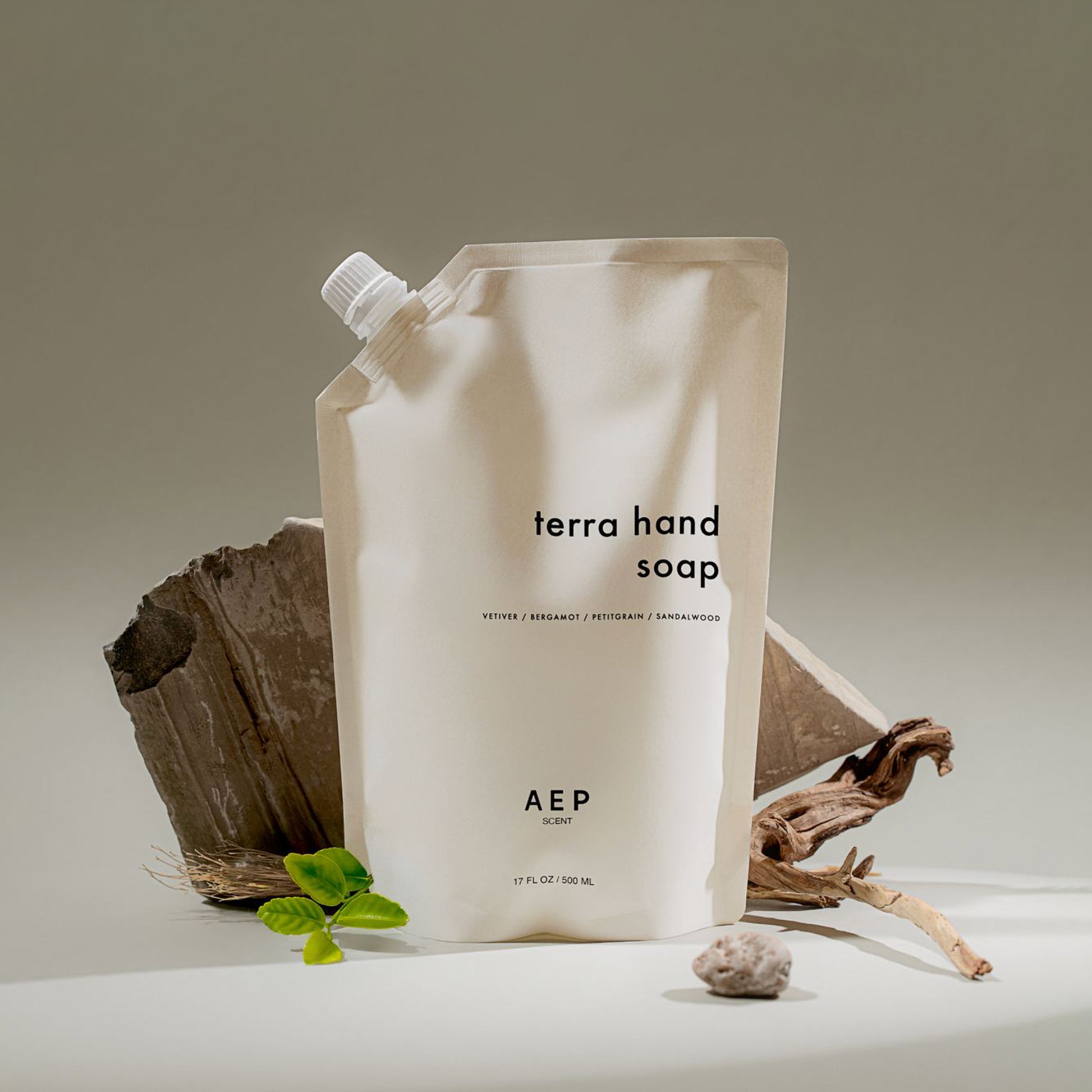 AEP Scent Terra hand soap refill pouch photographed with natural wood and green leaves, featuring notes of vetiver, bergamot, petitgrain, and sandalwood on a beige background.